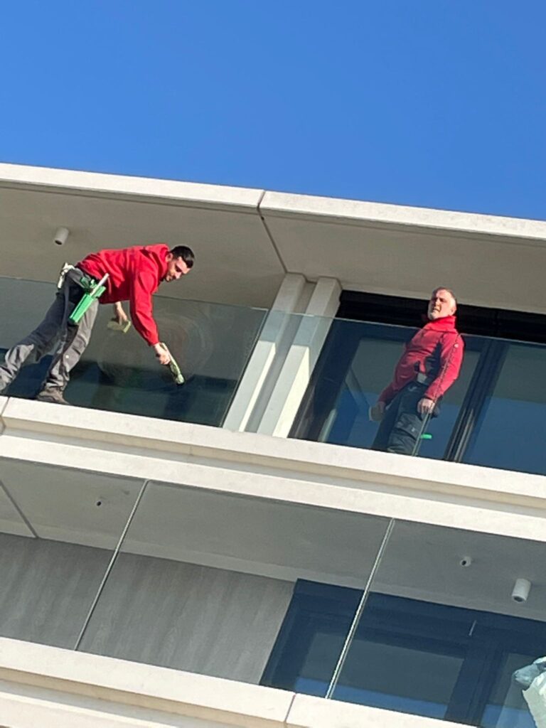 Twee glazenwassers schoonmaken balkonramen van een modern gebouw tegen een heldere blauwe lucht.
